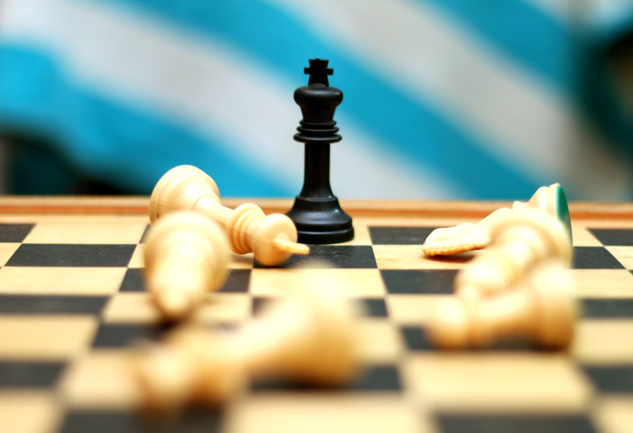 Close-up of a black king chess piece standing on a board surrounded by fallen white pieces.
