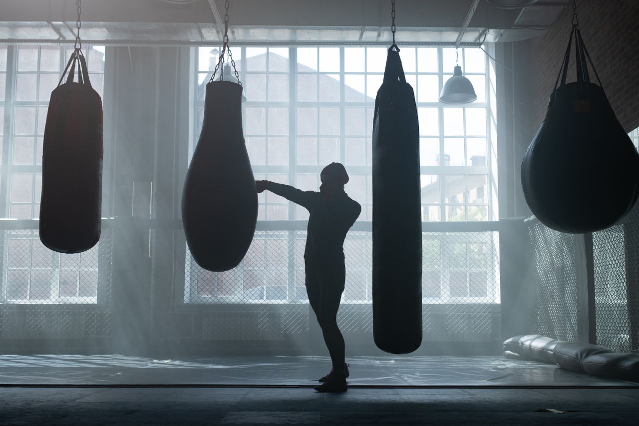 A silhouette of a person training with punching bags in an industrial-style boxing gym with large windows.